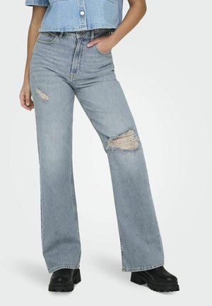 Jeans taille haute en denim bleu clair usé avec des déchirures, présentant une coupe large et des coutures contrastantes, assortis à des bottes épaisses à plateforme noires.