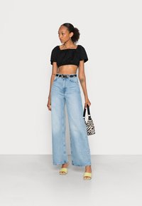 Crop top preto, jeans de perna larga de cintura alta azul clara, sapatos de salto amarelos de biqueira aberta, bolsa com padrão zebra e tatuagem visível no braço.