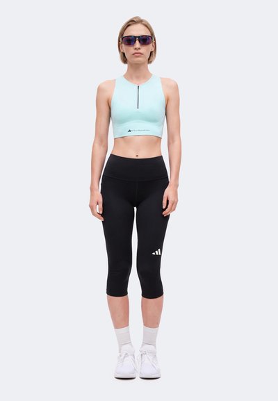 Frau trägt hellblaues ärmelloses Crop-Top, schwarze Capri-Leggings mit Logo, weiße Turnschuhe, Socken und dunkle Sportsonnenbrille, steht aufrecht.