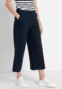 Donkerblauwe cropped broek met een brede pijp, een elastische tailleband en zijzakken, gecombineerd met witte sneakers op een effen achtergrond.