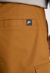 Barna vászonsort, amely texturált anyagból készült és rácsos mintázattal rendelkezik, fekete Nike logóval ellátott címkével a hátsó zsebében.
