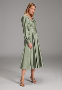 Robe à manches longues en satin vert clair en forme de A avec un décolleté en V et une taille froncée. Présente des manches bouffantes et une texture lisse. Talons argentés à brides.