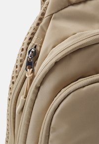 Beige ryggsäck med strukturerad tyg, en stor dragkedja och nätpaneler. Har rundade kanter och förstärkta sömmar för hållbarhet.