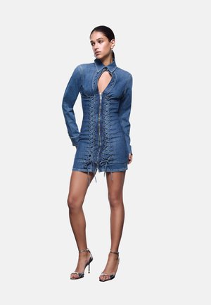 Donna che indossa un miniabito aderente in denim a manica lunga con dettagli frontali allacciati, una cerniera e un taglio sul petto, abbinato a sandali con tacchi alti.