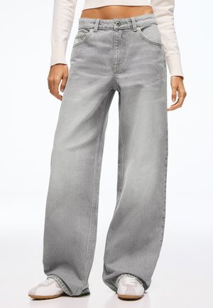 Femme portant un jean gris taille haute à jambes larges avec poches avant, associé à des baskets blanches et un top blanc court à manches longues.