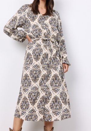 Kobieta ubrana w sukienkę z długim rękawem i paskiem, z misternym czarno-szaro-złotym kwiatowym wzorem na beżowym tle.