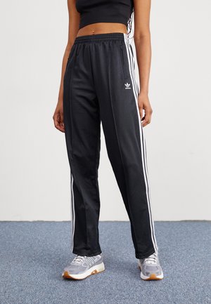 Person wearing black Adidas track pants with white stripes and gray sneakers, standing on blue carpet against white wall.