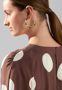 Boucles d'oreilles en or avec des perles colorées vertes et rouges. Blouse en tissu marron avec de grands pois blancs et des détails froncés aux épaules.