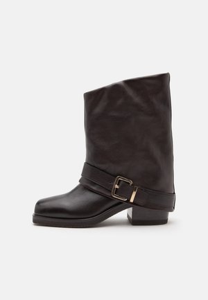 Bottine en cuir marron foncé avec un petit talon carré et une bride décorative avec une boucle dorée autour de la cheville.
