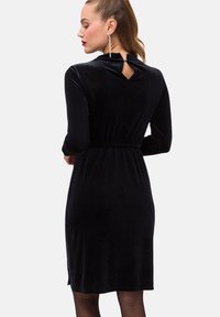 Czarna sukienka z weluru z długimi rękawami, dopasowaną talią, detalem w kształcie dziurki na plecach oraz gładką teksturą, sięgająca do połowy łydki.