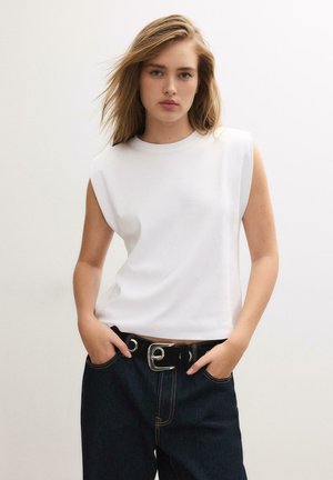 Jeune femme aux cheveux blonds portant un haut blanc sans manches et un jean foncé, les mains dans les poches, sur fond uni.