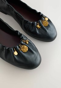 Fekete bőrből készült balettcipők, ráncolt szegéllyel, kis aranyozott medálokkal díszítve, mint csillag, kerek korongok és masni.