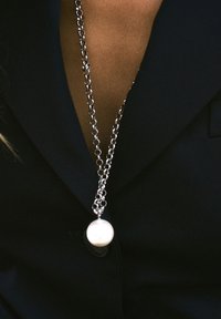 Hõbedane kettkaelakee, millel on suur ümmargune valge pärl ripats, mis puhkab tumeda kangaste tausta vastu.