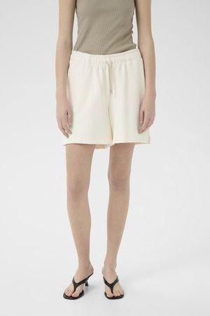 Lys kremfargede shorts med elastisk midjebånd og snor, med en avslappet passform og glat tekstur, brukt sammen med svarte flip-flops.