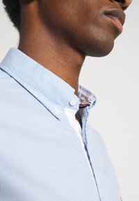 Gros plan d'une personne portant une chemise bleu clair à boutons, avec un col intérieur blanc et un détail rayé rouge et noir.
