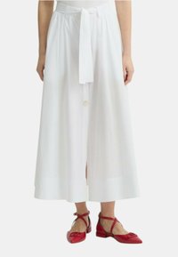 Gonna bianca midi con cintura a fiocco, chiusura con bottoni e spacco frontale; indossata con scarpe basse rosse con dettaglio a fiocco.