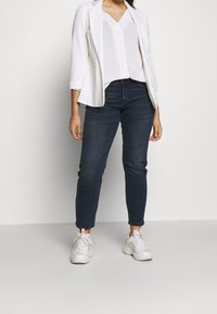 Bílý žebrovaný blejzr přes bílý top s výstřihem do V, doplněný tmavě modrými slim džíny a bílými teniskami. Jednoduchý design s neformálním vzhledem.