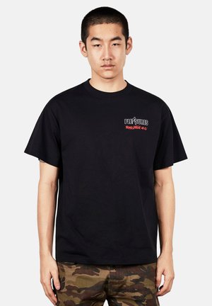 Mężczyzna z krótkimi włosami ma na sobie czarny t-shirt z napisem ""PLEASURES Too Hot 4 U"" i spodnie w moro, stoi na tle jednolitego tła.