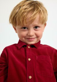 Lyshåret ung dreng smilende iført en rød kordfløjls-skjorte med knapper og en forlommer, mod en ensfarvet hvid baggrund.