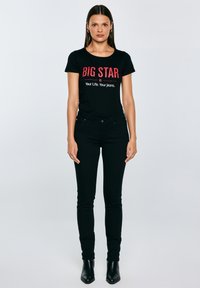 Egy nő szemben áll egy egyszerű világos háttér előtt, fekete "BIG STAR" feliratú pólót, fekete farmert és fekete bokacsizmát viselve.