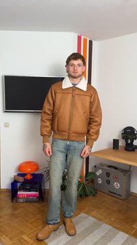 Giacca bomber in pelle marrone con texture, colletto in shearling bianco, chiusura a zip, polsini in maglia, abbinata a jeans azzurri chiari e stivali marroni.