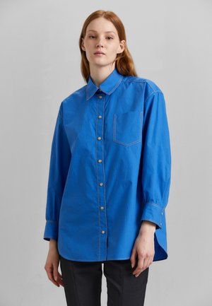 Blå skjorte med lange ermer, lys søm, enkelt brystlomme og buet skjortefyll. Glatt tekstur og skreddersydd passform.