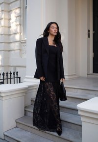 Juodas nėrinių suknelė, pasižyminti srauniu dizainu, sluoksniuota virš prigludusios juodos palaidinės. Derinama su pritaikytu juodu švarku ir mažomis rankinėmis.