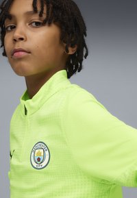Neon keltainen urheilu paita, jossa on teksturoitu kuvio, korkea kaulus ja Manchester Cityn logo rinnassa. Sisältää mustia yksityiskohtia ja vetoketjun.