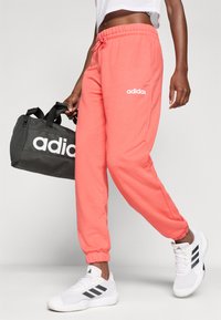 Korálové bavlněné tepláky mají elastický pás a manžety, s bílým logem adidas. V druhé ruce drží černou sportovní tašku adidas.
