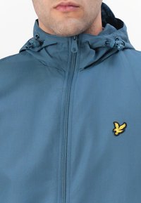 Modrá voděodolná bunda s předním zipem a nastavitelnou kapucí. Obsahuje žlutě vyšívané logo na levém prsu.