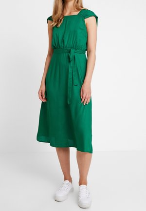 Mujer con un vestido midi verde sin mangas con cinturón en la cintura y zapatillas blancas de pie contra un fondo liso.