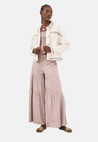 Chaqueta de mezclilla blanca con acentos bordados, combinada con una falda maxi de capas en rosa claro. La modelo lleva sandalias doradas y una blusa texturizada.