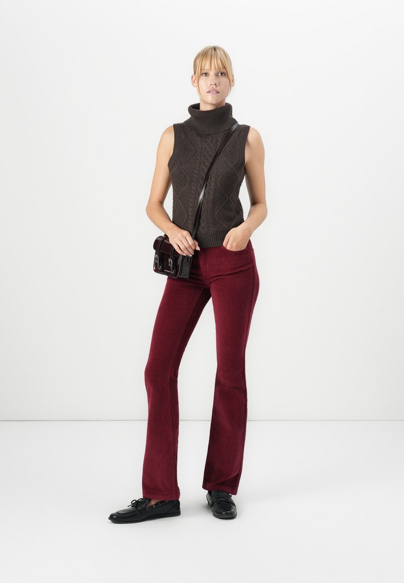 Femme portant un pull en col roulé sans manches marron, un pantalon en velours côtelé bordeaux, des chaussures noires et portant un petit sac à bandoulière noir.