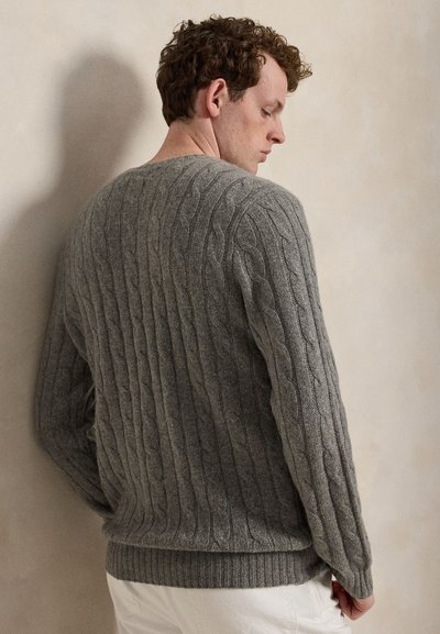 Mann mit lockigem Haar, der einen grauen Kabelstrickpullover und weiße Hosen trägt, steht mit dem Rücken zugewandt und leicht gesenktem Kopf.