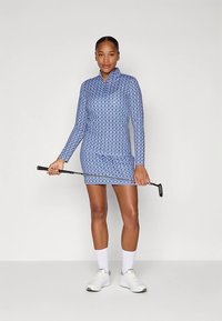 Kobieta w niebieskiej sukience do golfa z długim rękawem w wzory, trzymająca putter, ubrana w białe skarpetki i białe buty golfowe, stojąca na jednolitym tle.