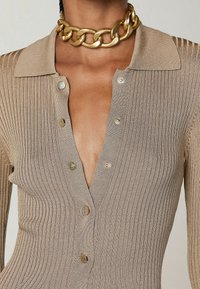 Haut en maille côtelée beige avec col, doté d'un profond décolleté en V, de boutons dorés et d'une coupe ajustée. Mis en valeur par un collier chaîne doré.