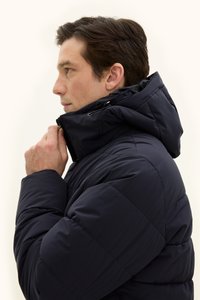 Navy-blauer Pufferjacke mit hohem Kragen und abnehmbarem Kapuzen. Verfügt über ein gestepptes Muster und einen Reißverschluss, gesehen aus einer Seitenansicht des Trägers.