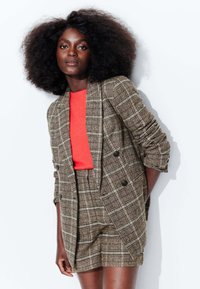 Blazer à carreaux brun double boutonnage avec un haut orange contrastant et un short à carreaux assorti. Texture douce et design structuré avec des manches retroussées.
