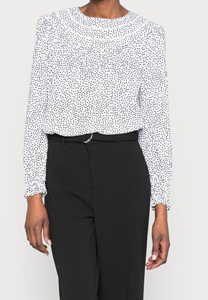 Kvinde iført en hvid langærmet bluse med sorte polkaprikker, stoppet ned i sorte bukser med høj talje og bælte.