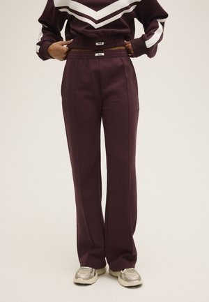 Persona que lleva pantalones anchos de pierna en color púrpura oscuro y sudadera a juego con rayas en forma de cheurón blancas, combinados con zapatillas beige.