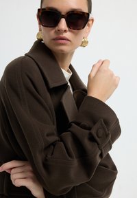 Bruine wollen jas met een brede kraag, oversized pasvorm en decoratieve knopen op de mouwen. Gecombineerd met oversized schildpadbruine zonnebril.