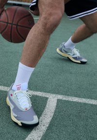 Szürke és fehér kosárlabda cipő, texturált fels�őrésszel, sárga talppal és piros márkajelzéssel, fehér sportzoknival viselve egy zöld pályán.