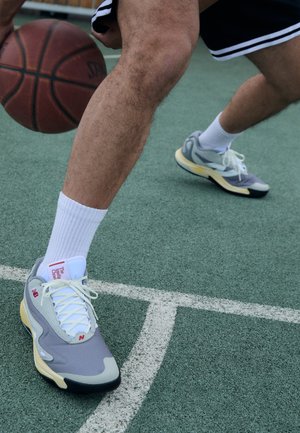 Sneaker de baschet gri și alb, cu un superior texturat, talpă galbenă și branding roșu, purtat cu șosete atletice albe pe un teren verde.