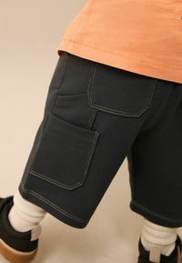 Donkergrijze shorts van katoen met twee grote zijzakken, afgewerkt met contrasterende gele stiksels. G gedragen met witte sokken en sneakers.