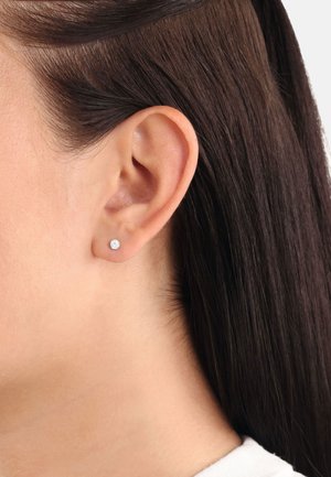 Boucle d'oreille ronde avec une pierre précieuse claire. Monture en métal, design minimaliste. Les cheveux sont coiffés en arrière, exposant la boucle d'oreille et l'oreille.