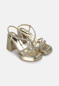 Gold, square-toed heeled sandals with a chunky block heel, featuring multiple crisscross straps adorned with iridescent accents.
