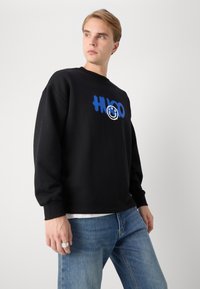 Crna dukserica s okruglim izrezom, dugim rukavima i plavim grafičkim dizajnom koji prikazuje smajlića i natpis. Uparena s svijetlo plavim trapericama.