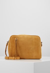 Bolso cruzado rectangular de ante color marrón con panel frontal perforado, bolsillo con cremallera vertical y correa de cuero ajustable sobre una superficie blanca.