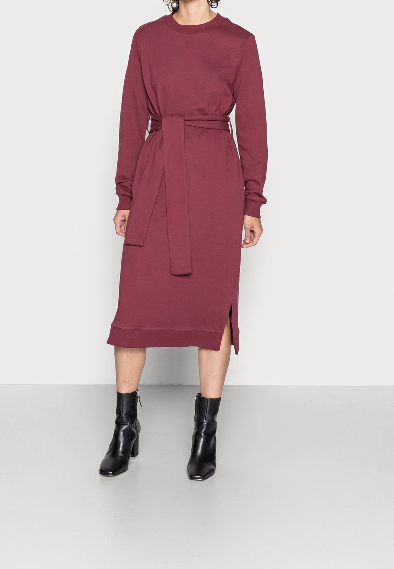 Femme portant une robe midi bordeaux à manches longues avec une ceinture à la taille et une fente latérale, associée à des bottines noires à talons.
