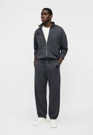 Homme debout les mains dans les poches, portant un survêtement zippé gris foncé et des baskets blanches sur un fond clair uni.